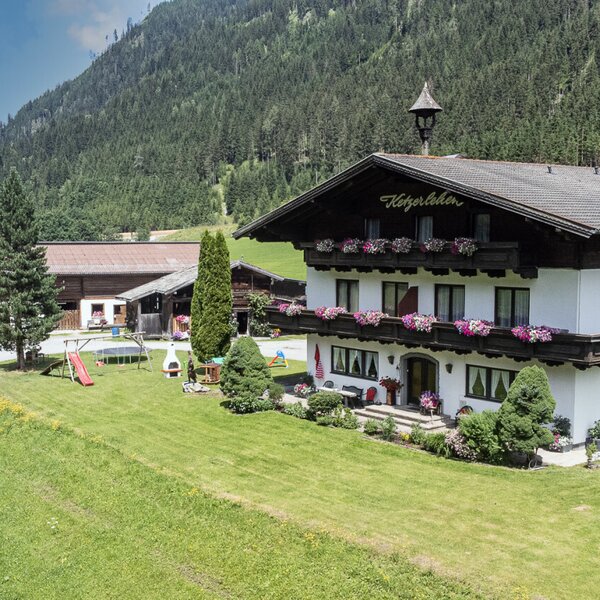 The Farm House exterior features flower-decorated balconies, a surrounding lawn, and a children's play area with a slide and trampoline, set against a forested mountain.