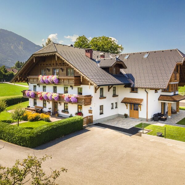Außenansicht des Bauernhofs mit blumengeschmückten Balkonen, einem Gartenbereich und Parkmöglichkeiten, umgeben von Wiesen und Bergen.