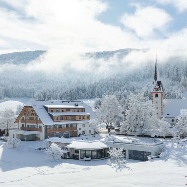 Der Bauernhof in einer verschneiten Winterlandschaft, umgeben von dem Dorf und den bewaldeten Bergen.