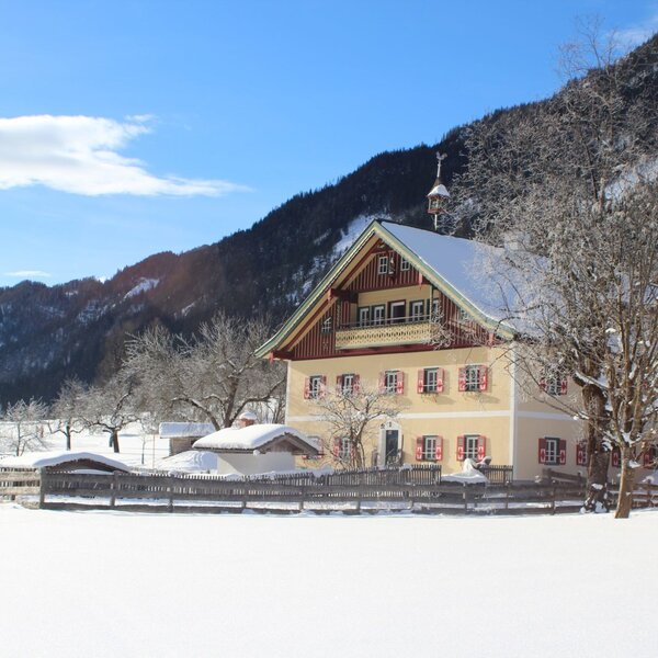 The yellow-painted Farm House with red shutters and a snow-covered roof, featuring an upper balcony, surrounded by snow and mountains.