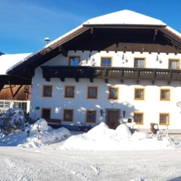 Das Äußere des Bauernhofs im Winter, umgeben von verschneiten Bäumen und Bergen unter blauem Himmel.
