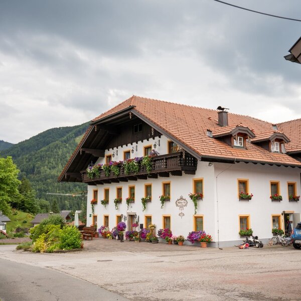 Der Bauernhof mit weißer Fassade, Holzbalkonen, Blumenkästen und Bergen im Hintergrund.