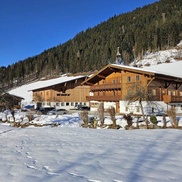 The Mayrhof farmhouse in winter, with its wooden buildings and balconies, set against a snow-covered, forested mountain.