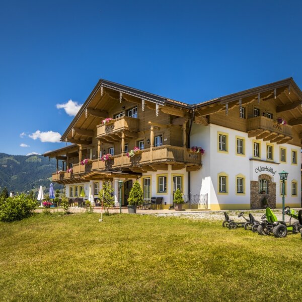 The Farm House exterior showcases wooden balconies with flower boxes, an outdoor terrace, a lawn with go-karts, and mountain views.