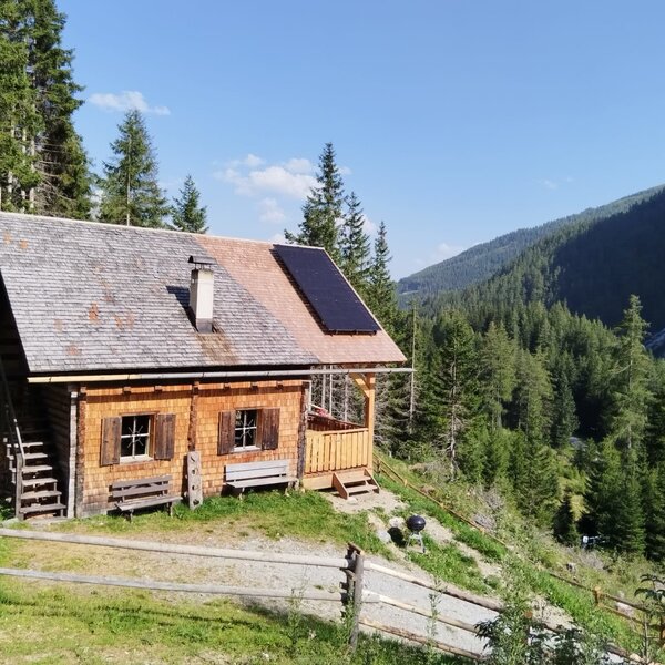 The wooden farmhouse features solar panels on the roof, outdoor benches, and a mountain forest setting.