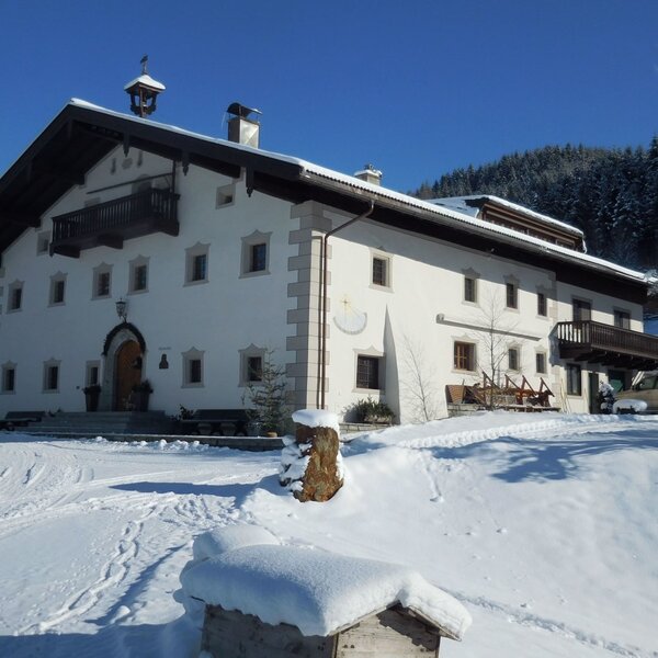 Der Bauernhof mit seiner weißen Fassade und den Holzbalkonen in der winterlichen, schneebedeckten Berglandschaft.