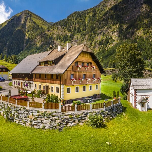 The Farm House, featuring a yellow facade, wooden upper levels with flower boxes on balconies, and an outdoor seating area, surrounded by green mountain slopes.