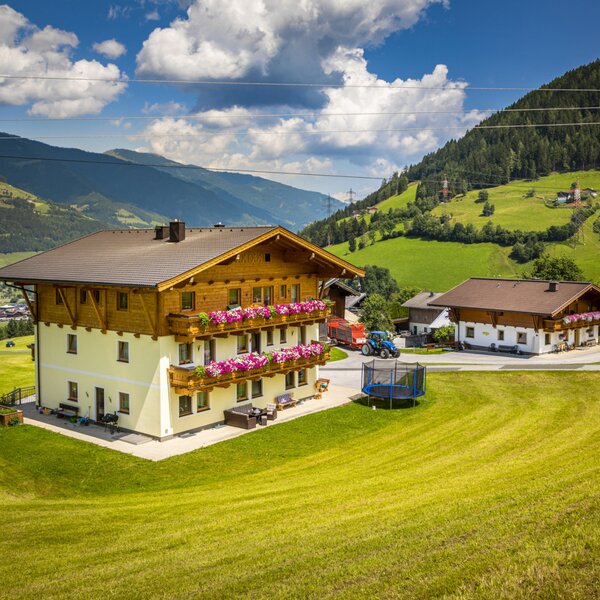 The main farmhouse building with flower-lined balconies and a trampoline, set amidst green fields with mountain views.