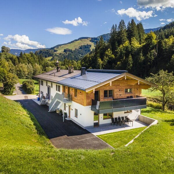 The farmhouse exterior, featuring wood and white finishes, balconies, and an outdoor patio with seating, is situated amidst green hills and mountains.