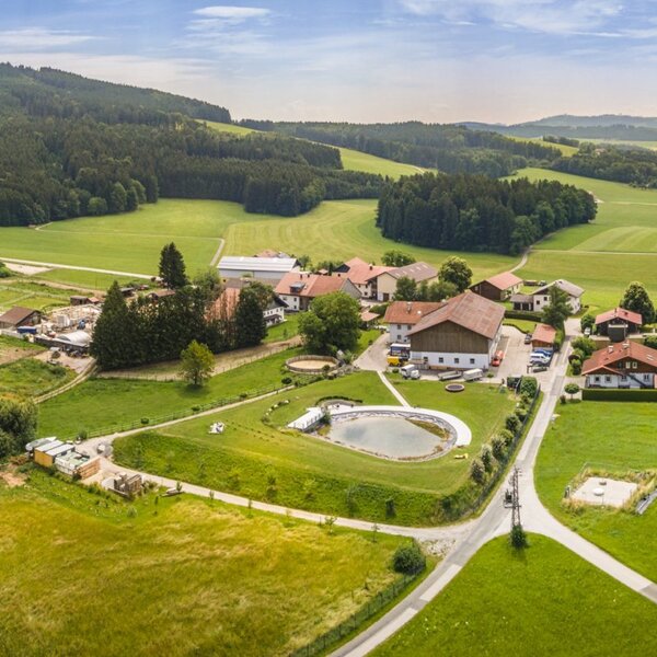 Der Bauernhof aus der Vogelperspektive, eingebettet in grüne Felder und Wälder, mit einem Naturbadeteich auf dem Gelände.