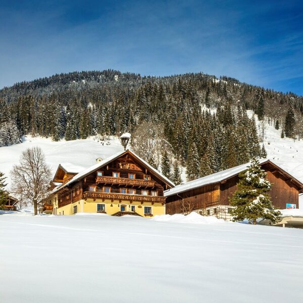 Der Bauernhof im Winter, umgeben von schneebedeckten Feldern und Wäldern vor einer Bergkulisse.