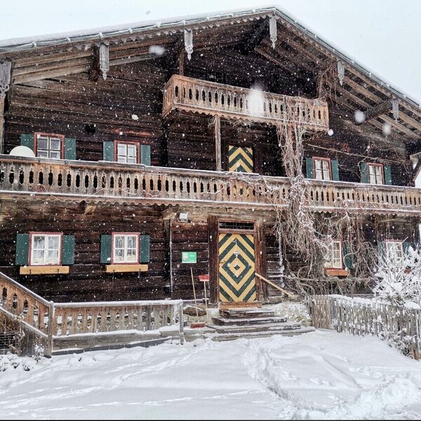 The wooden farmhouse exterior, featuring balconies with ornate railings, green-shuttered windows, and a distinctive patterned main door, pictured in the snow.
