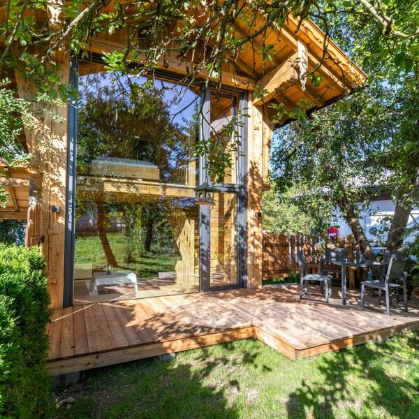 The Farmhouse provides a wooden deck with outdoor dining furniture, surrounded by a garden, next to a glass-fronted section of the building.