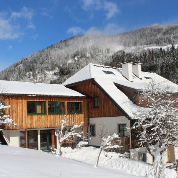The exterior of the Farm House, featuring snow-covered roofs and wooden facades, nestled in a winter landscape with snow-capped mountains.