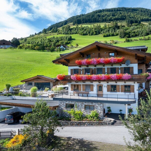 Außenansicht des Bauernhofs mit blumengeschmückten Balkonen, Carport und grüner Hügellandschaft.