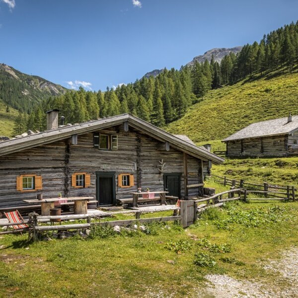 The farmhouse features an outdoor seating area with wooden tables and benches, surrounded by green mountain slopes, a wooden fence, and a nearby stream.
