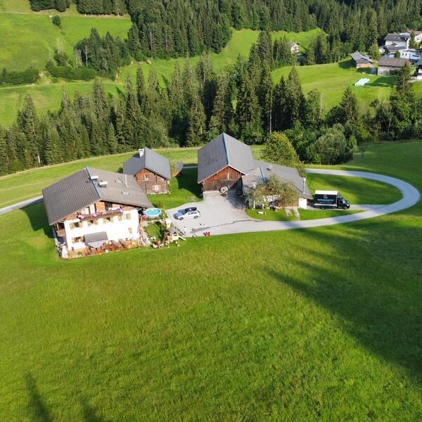 Luftaufnahme vom Bauernhof mit mehreren Gebäuden, umgeben von grünen Hügeln und Wäldern, mit einer Zufahrtsstraße.