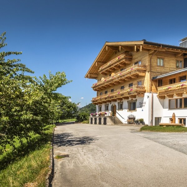 Der Bauernhof mit seiner traditionellen Holzarchitektur, Balkonen, einem angrenzenden Wirtschaftsgebäude und einer gepflasterten Zufahrt, umgeben von grüner Natur.