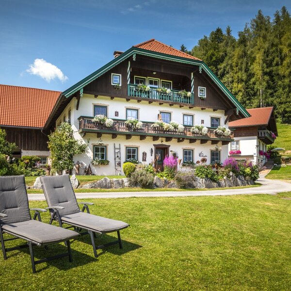 Der Bauernhof mit blumengeschmückten Balkonen und einer Liegewiese mit zwei Sonnenliegen vor einem bewaldeten Hügel.