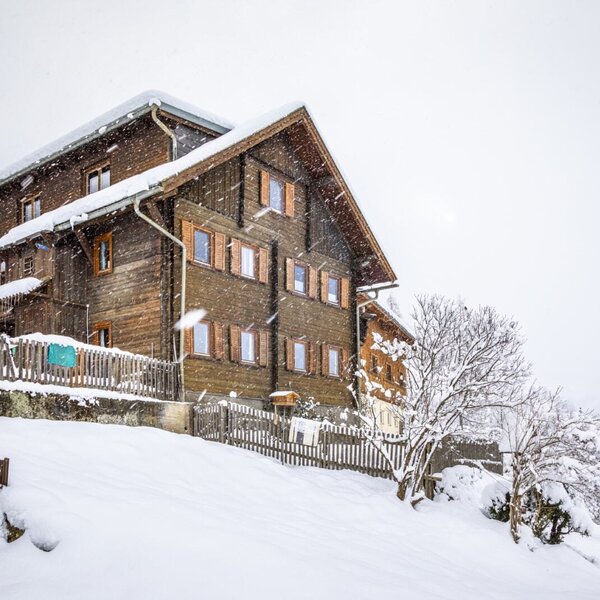 The wooden exterior of the farmhouse features multiple stories and shuttered windows, surrounded by a snow-covered landscape.