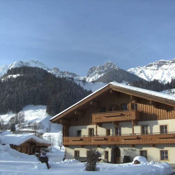 Außenansicht des Bauernhofs „Zieferhof“ mit schneebedecktem Dach und den verschneiten Bergen im Hintergrund.