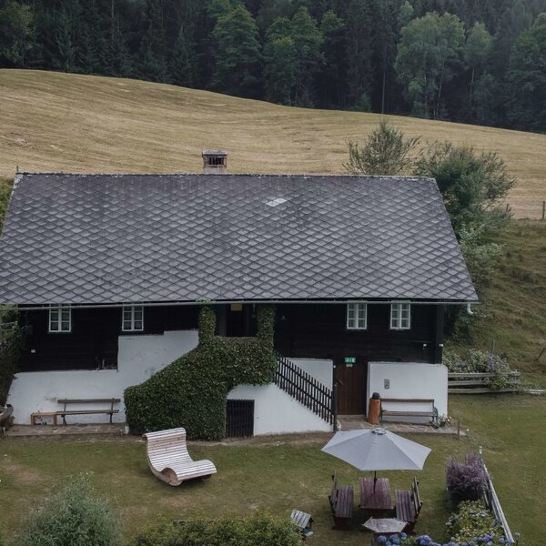 The farmhouse exterior includes a garden with an outdoor dining area, loungers, and benches, set against a backdrop of hills and forest.