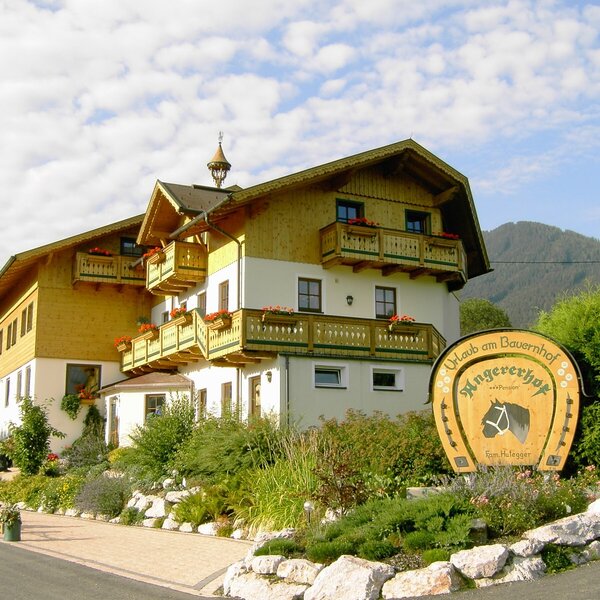 Die Außenansicht des Bauernhofs mit Holzbalkonen, Blumenkästen und einem Hinweisschild "Urlaub am Bauernhof Angererhof" im Vorgarten, mit Bergen im Hintergrund.