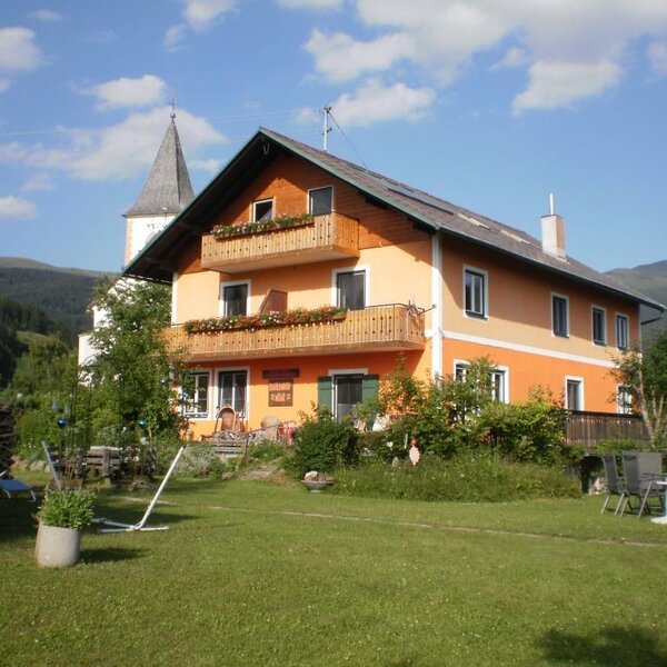 Der Bauernhof mit blumengeschmückten Balkonen, einer Rasenfläche und Blick auf die umliegenden Berge und den Kirchturm.