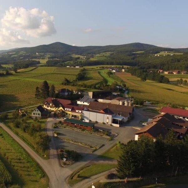 Aerial view of the farmhouse property, including its buildings and parking, set amidst green fields with hills in the background.