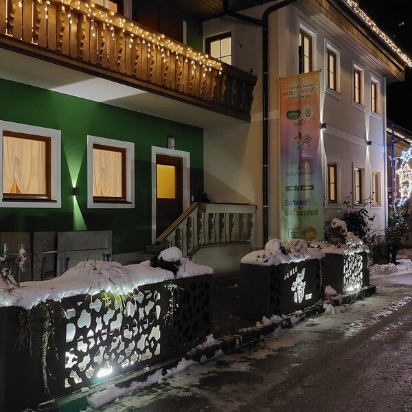Die beleuchtete Fassade des Bauernhofs bei Nacht, mit Lichterketten auf dem Balkon und weiteren festlichen Dekorationen im Schnee.