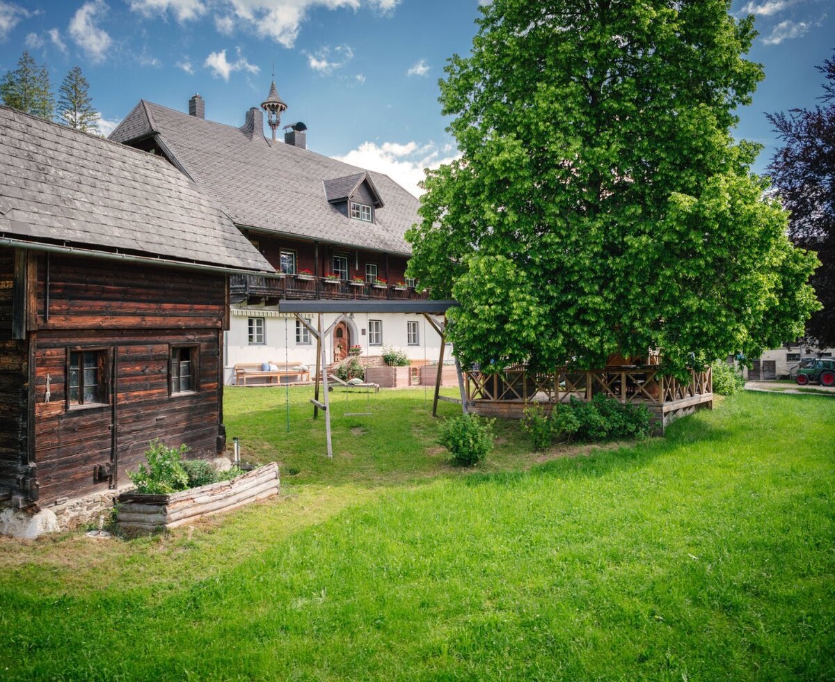 Der Bauernhof mit einem traditionellen Holzgebäude, einem großen Garten mit Schaukel und einem Baum mit Sitzbereich.