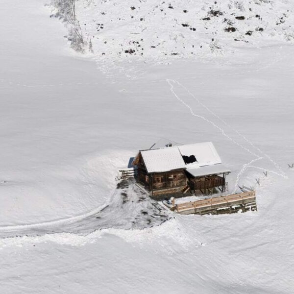 The wooden farmhouse, featuring a snow-covered roof, is situated in a winter landscape with an access path leading to the entrance.