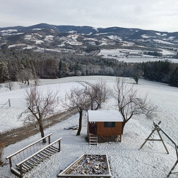Der Außenbereich des Bauernhofs im Winter mit Spielhaus, Schaukel, Feuerstelle und Panoramablick auf die schneebedeckten Hügel.