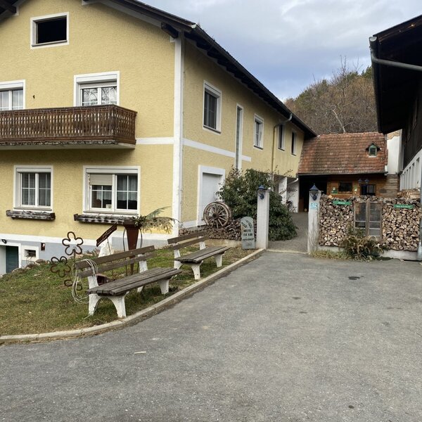 Die Außenansicht des Bauernhofs mit dem Hauptgebäude, einem Balkon und Sitzbänken am Wegesrand.