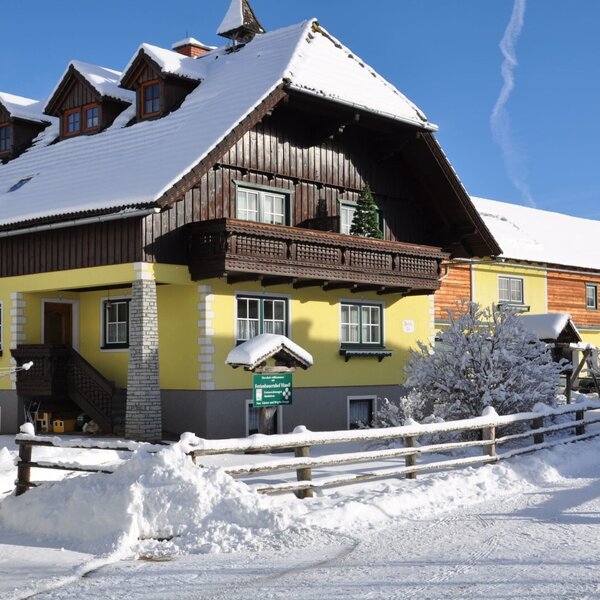 The farmhouse exterior showcases yellow walls and dark wooden accents, with a snow-covered roof and balconies during winter.
