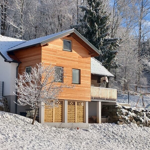 The two-story farm house, with wood and white cladding and a balcony, is situated in a snowy landscape with a forest background and a children's play structure.