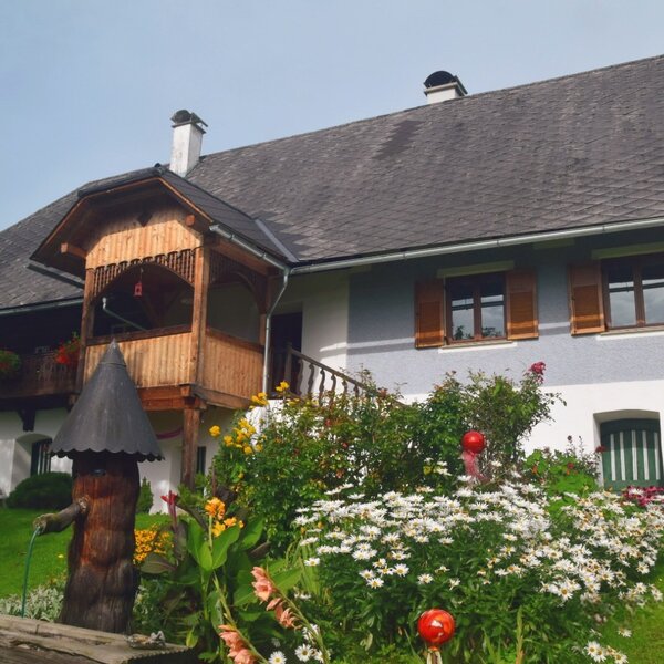 The farm house features a light blue and white exterior, a wooden balcony, and a front garden with a variety of flowers and a decorative wooden fountain.