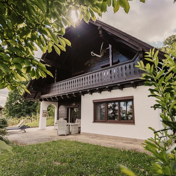 The farm house features a wooden balcony, a ground-floor patio with outdoor seating, and a surrounding lawn bordered by greenery.