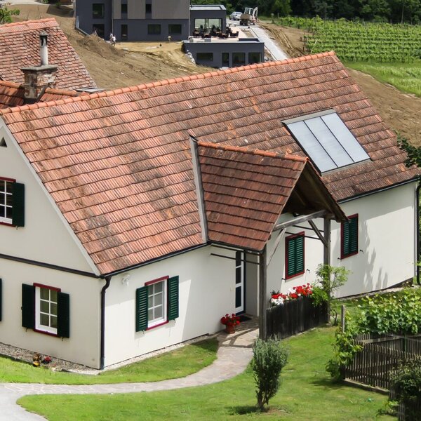 The farmhouse exterior features a red tiled roof, green window shutters, and roof-mounted solar panels, with a paved path flanked by flowers leading to the main entrance.