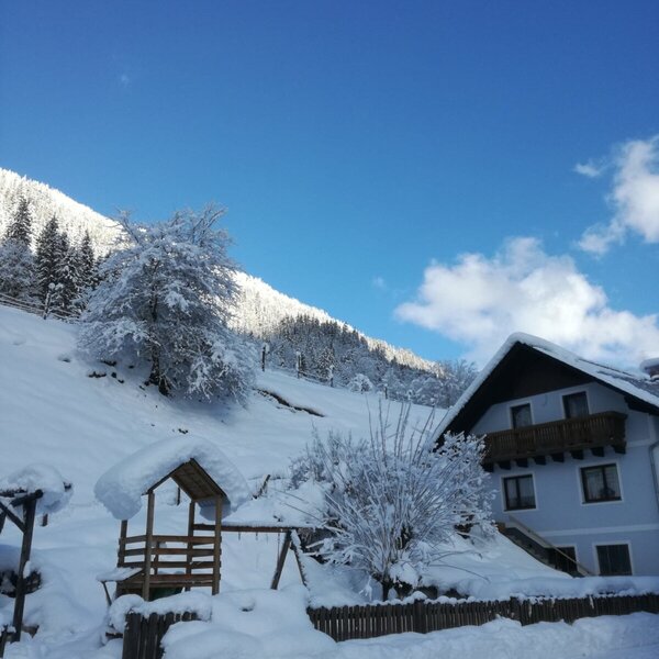 Der Bauernhof in winterlicher Umgebung mit schneebedecktem Spielplatz und Blick auf die Berge.