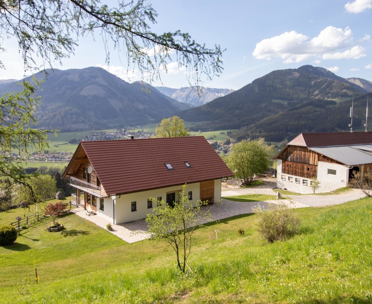 The farmhouse with a balcony and an adjacent farm building, situated on a green hillside with views of the surrounding mountains and valley.