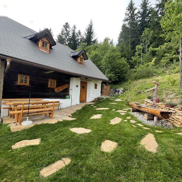 The farmhouse exterior includes an outdoor seating area with a wooden table and benches, a green lawn with stepping stones, and a decorative wooden water feature, surrounded by a forest.