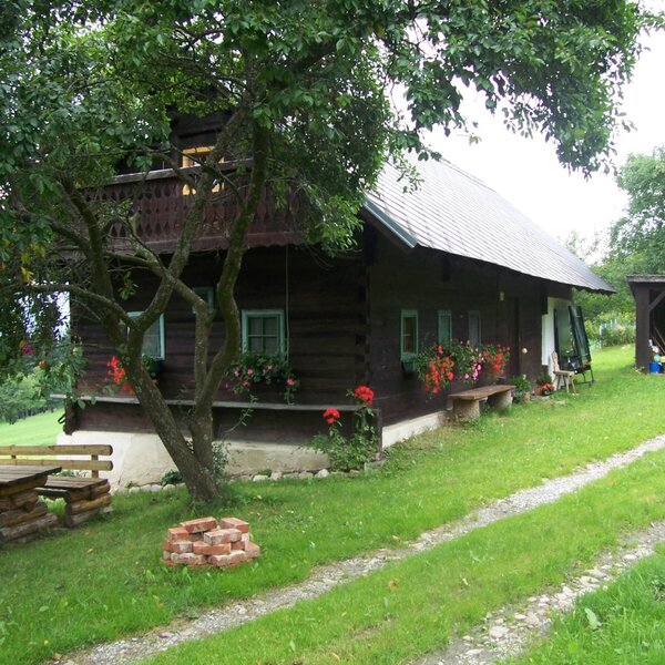 Die Außenansicht des Bauernhofs mit dunkler Holzfassade, Balkon, Blumenkästen an den Fenstern und einer Sitzgruppe aus Holz im Freien.