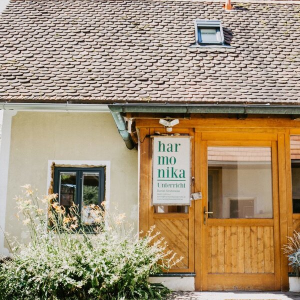 Die Außenansicht des Bauernhofs mit Ziegeldach, einem hölzernen Eingangsbereich und grünen Pflanzen vor den Fenstern.