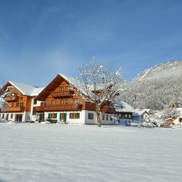 Der schneebedeckte Bauernhof mit traditionellen Holzbalkonen und der Bergkulisse im Hintergrund.
