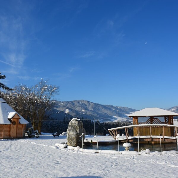 The Farm House grounds feature a snow-covered wooden hut and a gazebo with a bridge over a pond, set against a backdrop of snowy mountains.