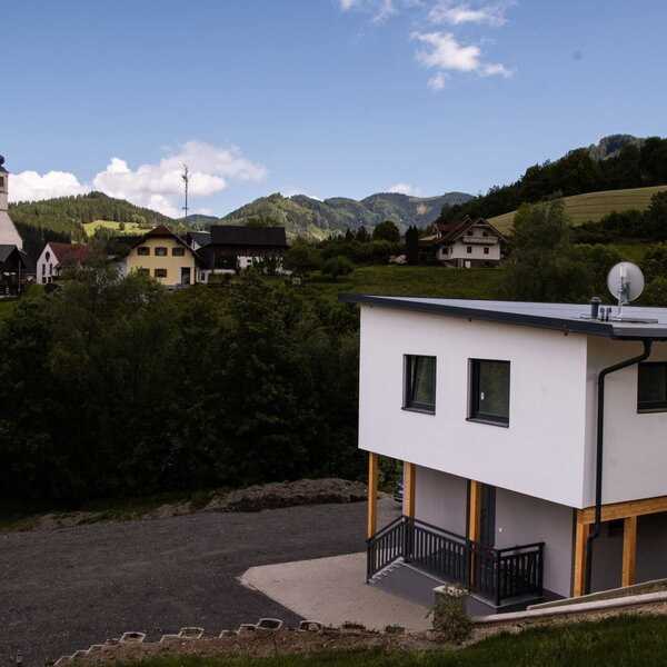 The exterior of the farm house features a white facade and dark windows, with a gravel parking area and views of the village and surrounding mountains.