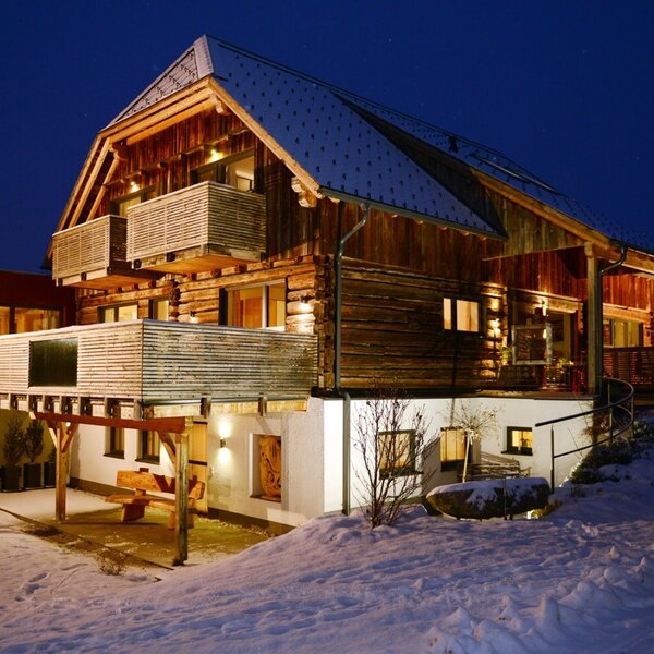 Der Bauernhof bei Nacht, mit Holzfassade, beleuchteten Balkonen und verschneiter Umgebung.