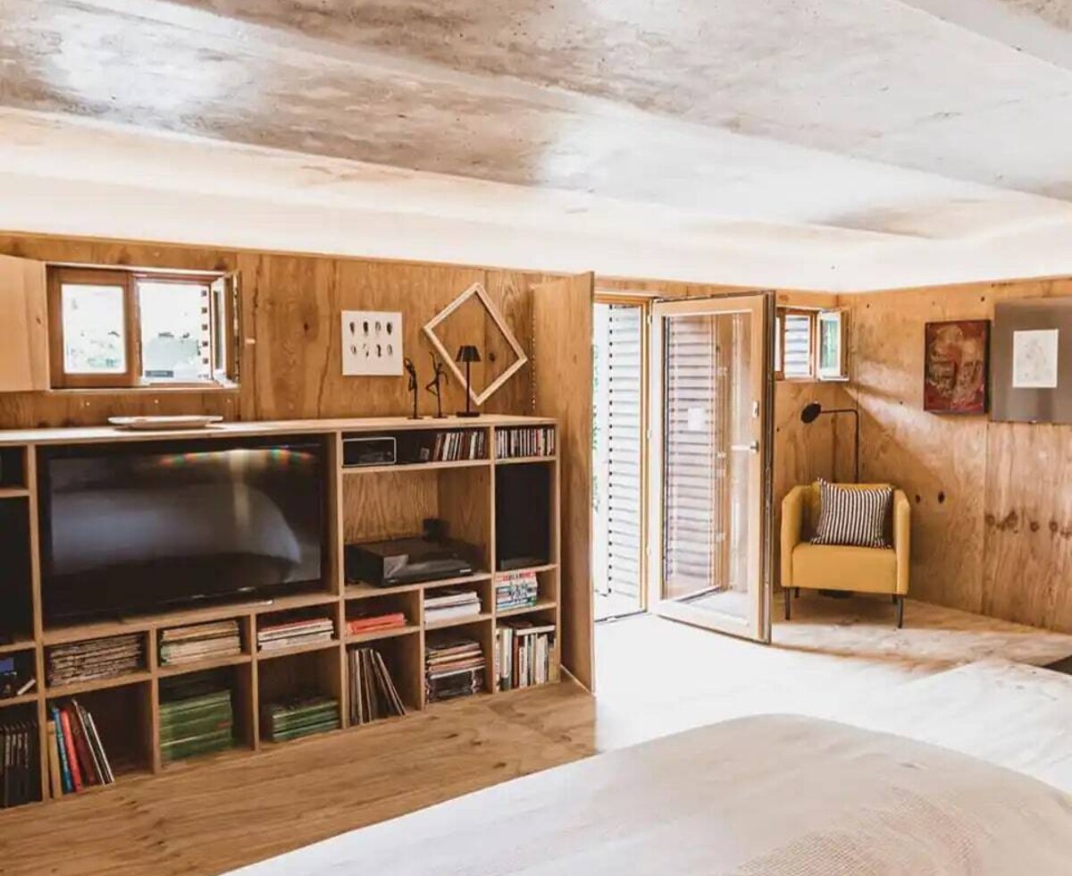 Schlafzimmer im Bauernhof mit großem Bett, Holzwänden, einem Regal mit Fernseher und Büchern, einem gelben Sessel und Zugang nach draußen.