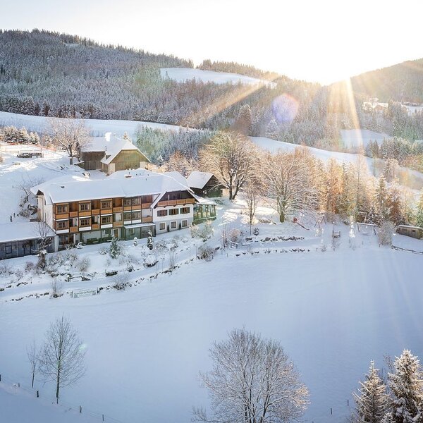 Der Bauernhof eingebettet in eine weitläufige, schneebedeckte Winterlandschaft mit Hügeln und Bäumen, unter strahlender Sonne.
